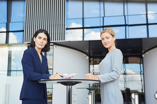Side view portrait of two businesswomen smiling at camera while standing by cafe table in airport or office building, copy space