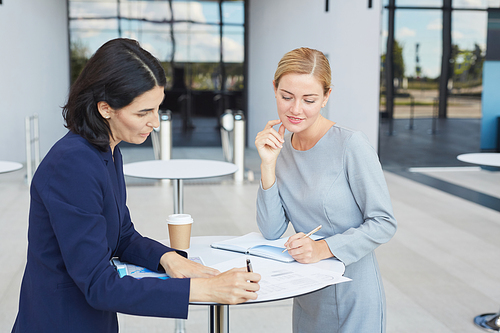Waist up portrait of two successful businesswomen discussing deal while standing by cafe table in airport or office building