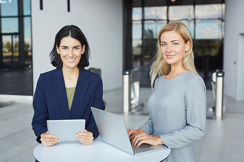 Waist up portrait of two successful businesswomen smiling at camera while standing at desk in office building and holding laptop, copy space