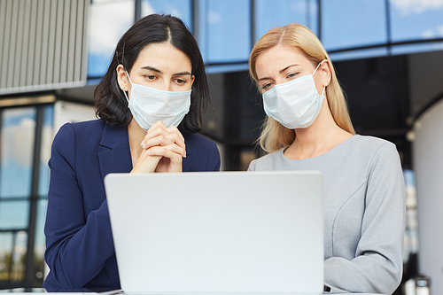 Waist up portrait of two successful businesswomen wearing masks while looking at laptop screen standing at desk in office building