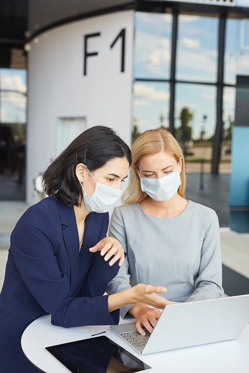 Vertical waist up portrait of two successful businesswomen wearing masks while looking at laptop screen standing at desk in office building