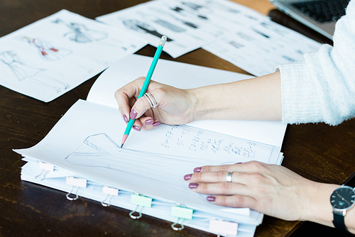 Hands of young female drawing fashion sketch on blank page of notepad for new seasonal collection while sitting by table