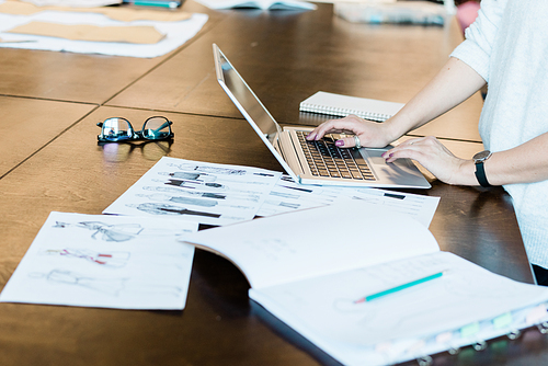 Close-up of unrecognizable woman standing at table with fashion sketches and using laptop in atelier