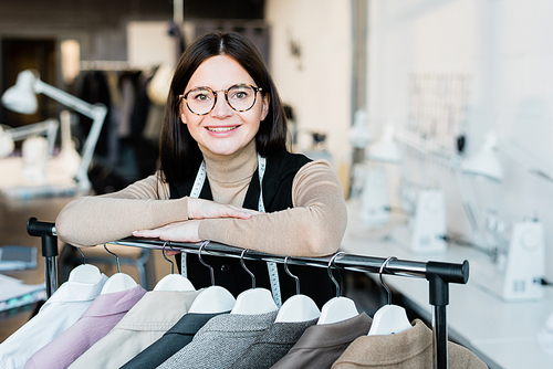 Happy young successful female fashion designer or shop assistant looking at you while standing by racket with new jackets