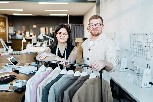 Team of young tailors in glasses standing at clothes rack in workshop and looking at camera