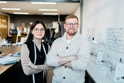 Two young contemporary fashion designers or tailors crossing their arms by chest while standing in front of camera in workshop