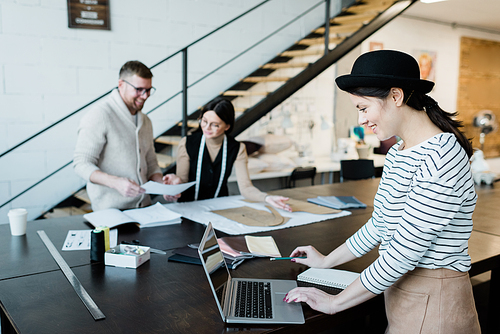 Happy young stylish woman in hat and casualwear looking at laptop display while standing by table in workshop