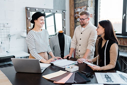 Three young happy designers discussing new fashion models for seasonal collection while working in studio