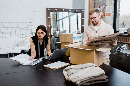 young man putting folded pants into carton box while packing parcel for one of clients while his colleague drawing sketch