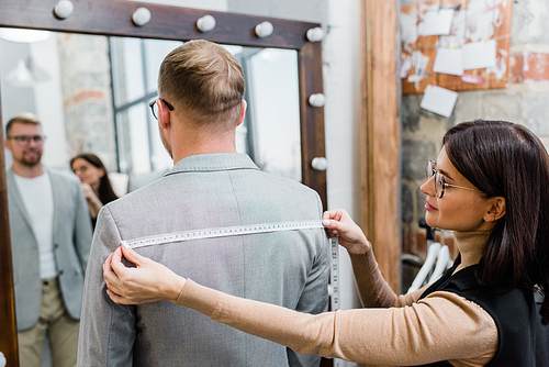 Rear view of female tailor measuring width of clients shoulders in own workshop