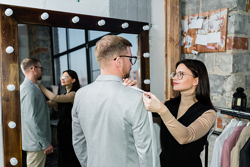 Young female tailor taking measures of jacket of young man while both standing by mirror in workshop or fashion studio