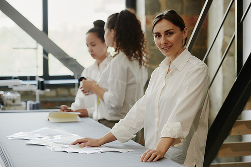 Young creative female designer of clothes looking at you while standing by table on background of two colleagues discussing new sketches