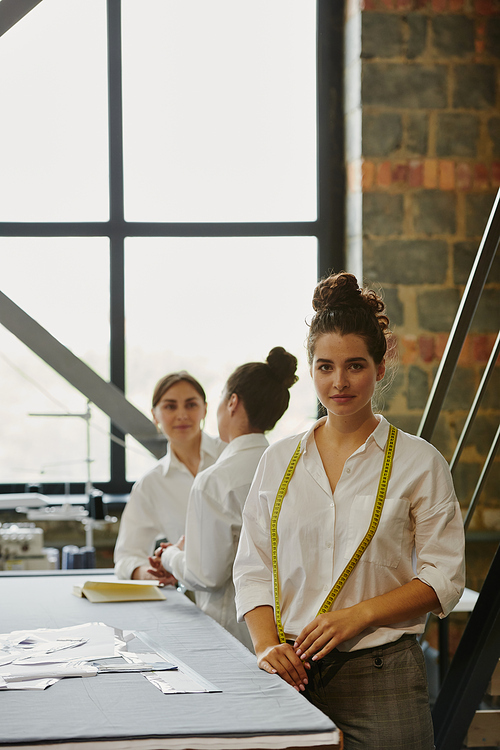 Young serious seamstress with measuring tape on her neck standing by table in workshop with her colleagues discussing ideas near by