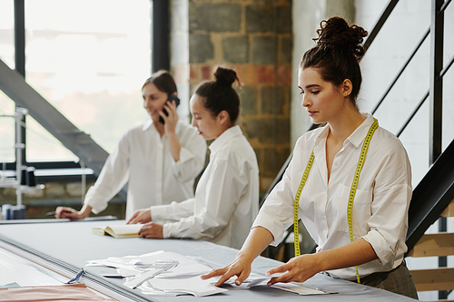 Young woman with measuring tape taking paper pattern from table while going to work over item of new fashion collection