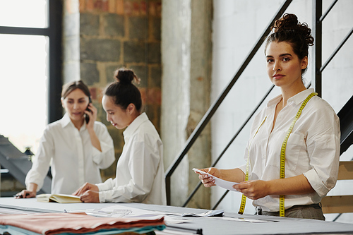 Young serious seamstress with measuring tape and paper patterns looking at you in workshop with her colleagues discussing ideas near by