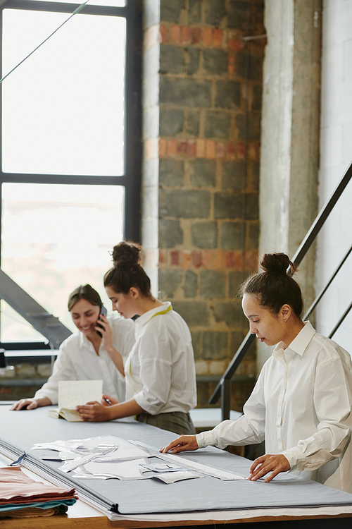 Two young designers of clothes phoning one of clients about order while their colleague measuring length of paper patterns near by