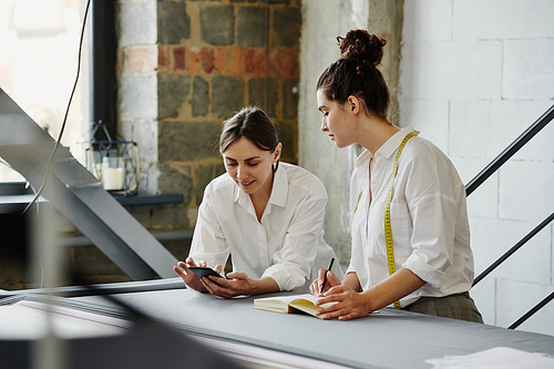 Young woman with mobile phone looking through online fashion trends while her colleague with notebook and pen making notes