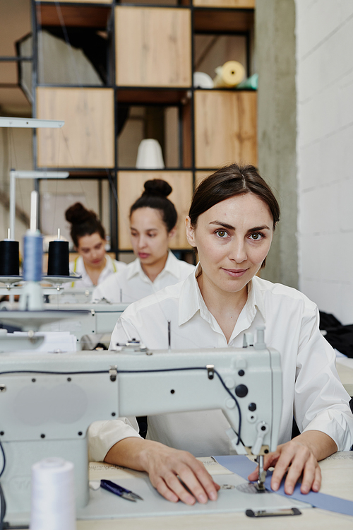 Young pretty seamstress in workwear looking at you while sitting by electric sewing machine and making or repairing clothes