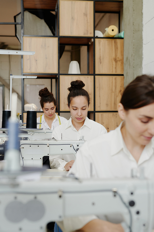 Contemporary seamstress in white shirt sitting by workplace with electric sewing machine and making or repairing clothes between colleagues