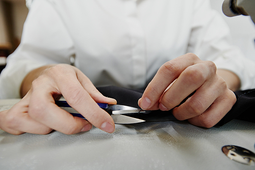 Hands of young contemporary seamstress or female tailor with sharp scissors cutting edges of black textile or thread by workplace