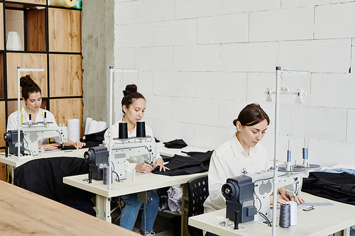 Row of young seamstresses sitting by desks and using electric sewing machines while working over new seasonal collection