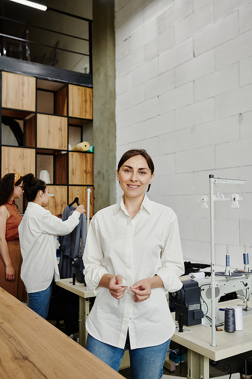 Young creative fashion designer in jeans and white shirt standing in front of camera in workshop on background of her two colleagues working