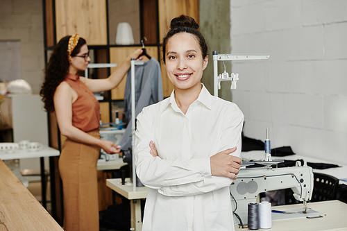 Young successful smiling seamstress crossing her arms on chest while standing in workshop on background of client with cardigan