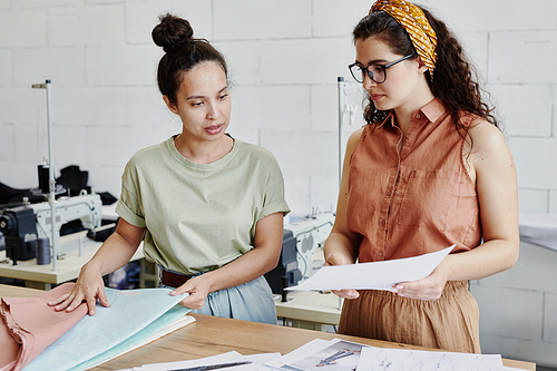 Young seamstress looking at fashion sketch on paper held by her colleague while choosing textile for new seasonal collection of clothes