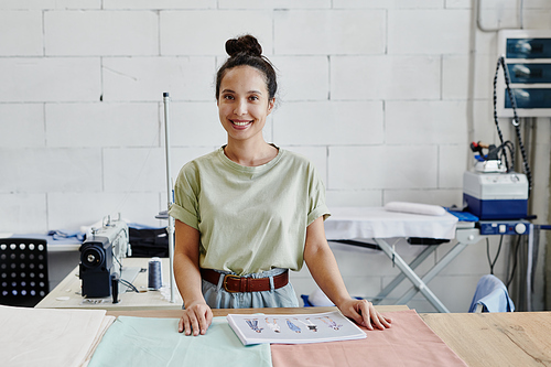 Young creative fashion designer looking at you while working with sketches for new seasonal collection of clothes while standing by table