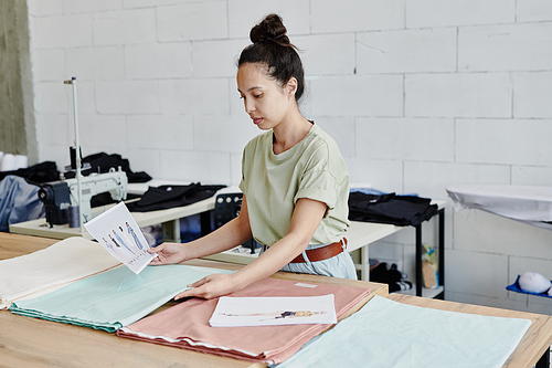 Young creative fashion designer looking for suitable textile for new seasonal collection of clothes while standing by table