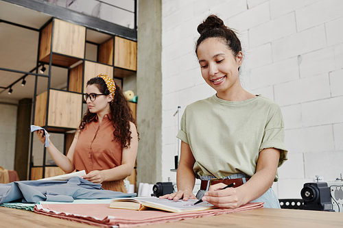 Happy young fashion designer pointing at sketch for new seasonal collection of clothes or drawing it while standing by table