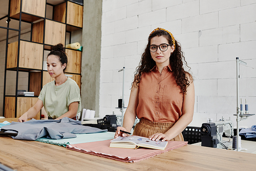 Young creative tailor or fashion designer in smart casualwear looking at you while standing by table with textile in workshop