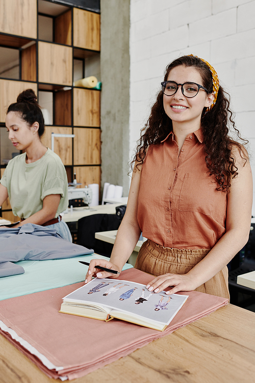 Young cheerful tailor or fashion designer in smart casualwear looking at you while standing by table in workshop and making sketches