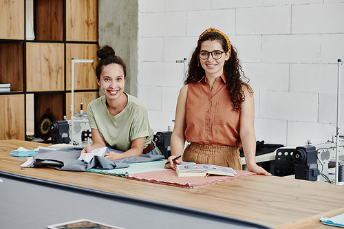 Two young successful fashion designers looking at you with smiles while choosing textile for new trendy fashion collection items