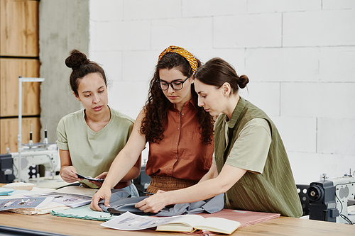 Three young fashion designers standing by table in workshop and choosing textile for one of items of seasonal collection