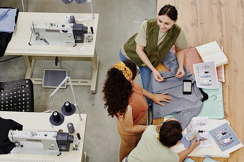 Group of contemporary seamstresses standing between table and sewing machines during discussion of new fashion collection in workshop