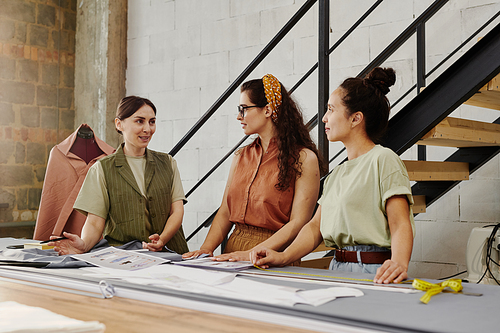 Two young female trainees showing tailoring coach new sketches of fashion items and consulting with her at start-up meeting