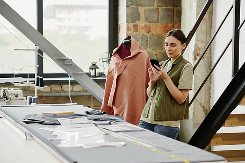 Contemporary young designer of fashion collection scrolling in smartphone through online ideas and new trends while standing by workplace