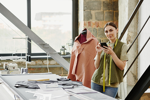 Happy young tailor and owner of fashion studio taking new orders from clients while standing by table with sketches and unfinished clothes