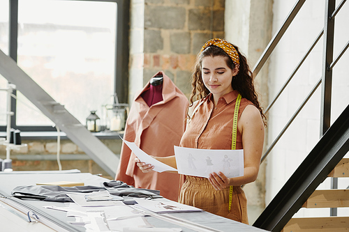 Young pretty fashion designer looking at sketches of new models for seasonal collection and choosing some of them for sewing