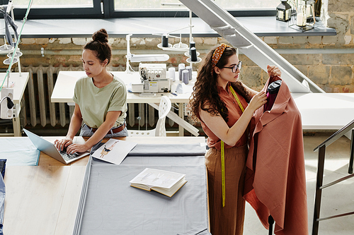 Two young serious fashion designers working over new seasonal collection of coats in studio while browsing in the net and working by dummy