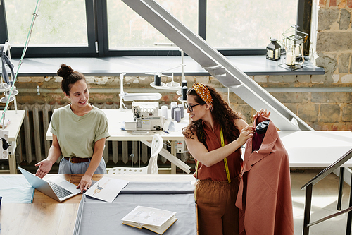 Happy young designer of fashion showing her colleague new models or items on laptop display while consulting with her about the choice