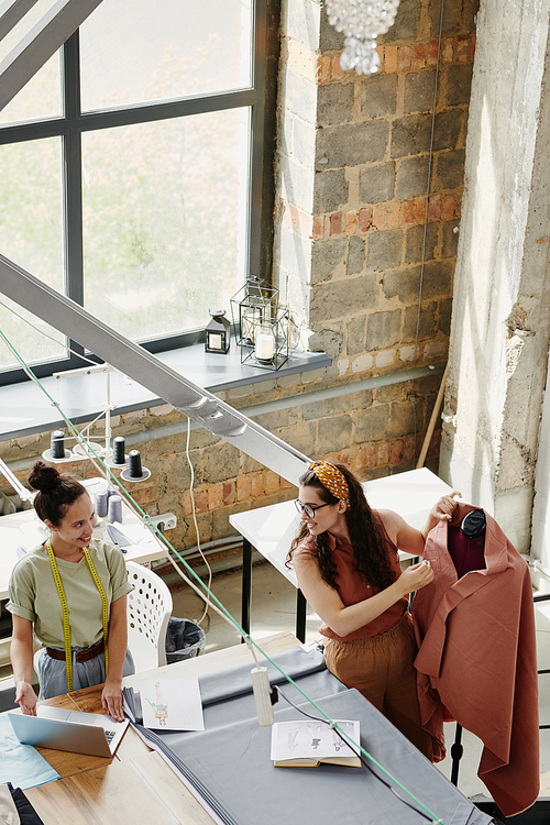 Happy young creative woman showing her colleague new models or items on laptop display while consulting with her about the choice