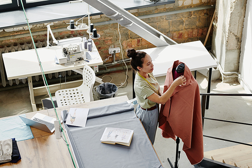 Young fashion designer working with textile on dummy while standing by table with fabric, laptop, open notebook and sketch