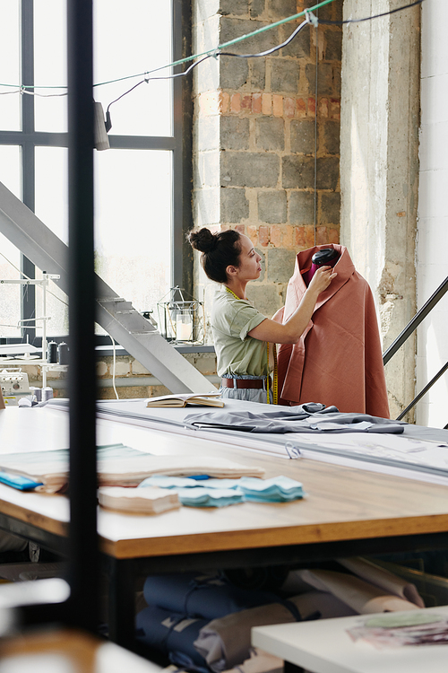 Female worker of modern fashion design studio sewing clothing item on dummy while working over new seasonal collection in workshop