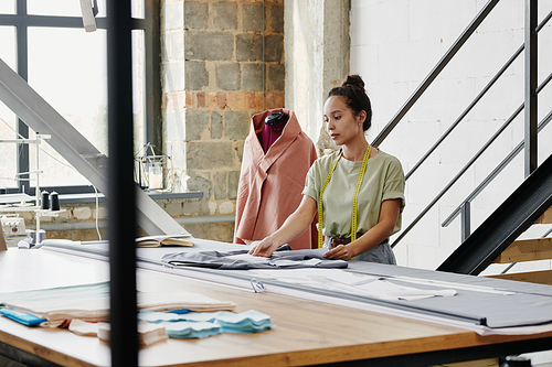 Young pretty designer of clothes in casualwear standing by large table in her studio or workshop and looking through new sketches