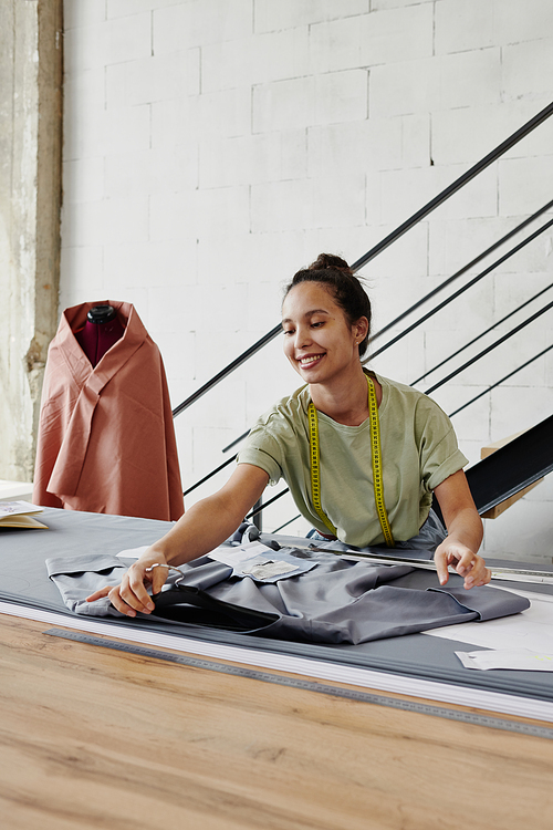 Pretty young smiling tailor working over new order of client or her own fashion collection while putting grey dress on table in studio