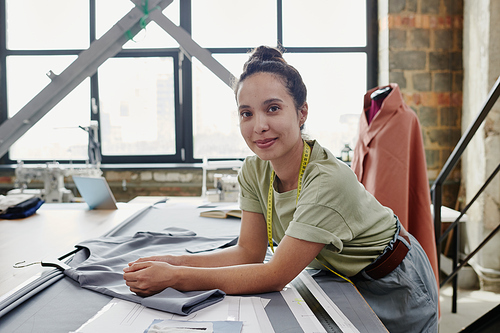 Young brunette tailor or designer of clothes leaning on table with unfinished dress, textile, paper patterns and ruler during work