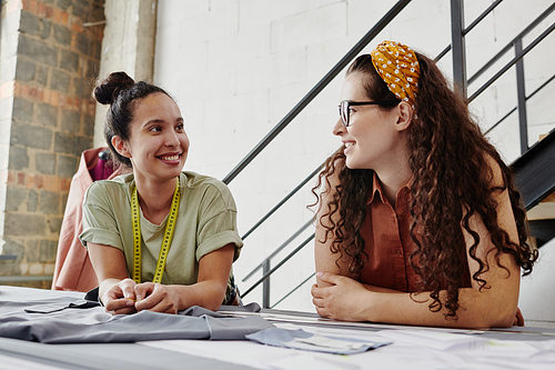 Young cheerful seamstress with measuring tape looking at colleague standing next to her during discussion of working moments