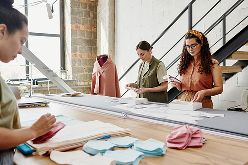 Group of three young designers of clothes standing by large table in workshop and looking through samples of fabric and other stuff